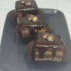 Close-up of three individual chocolate cakes with layers of cake and filling, topped with a glossy chocolate ganache and decorative chocolate pieces, arranged on a dark gray plate against a light gray background.