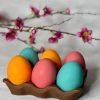 Six colorful, edible Easter eggs (teal, orange, pink) with a chocolate glaze, resting in a Dubai Chocolate style edible carton. The eggs contain Joconde sponge, mango and passion fruit insertion, and vanilla ganache. Pink blossoms are in the blurred background.
