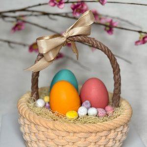A detailed view of a cake shaped like a woven Easter basket, containing large teal, orange, and pink frosted eggs, surrounded by smaller speckled and solid-colored candy eggs. A shiny gold bow is tied to the basket handle, and blurred pink blossoms are in the background.