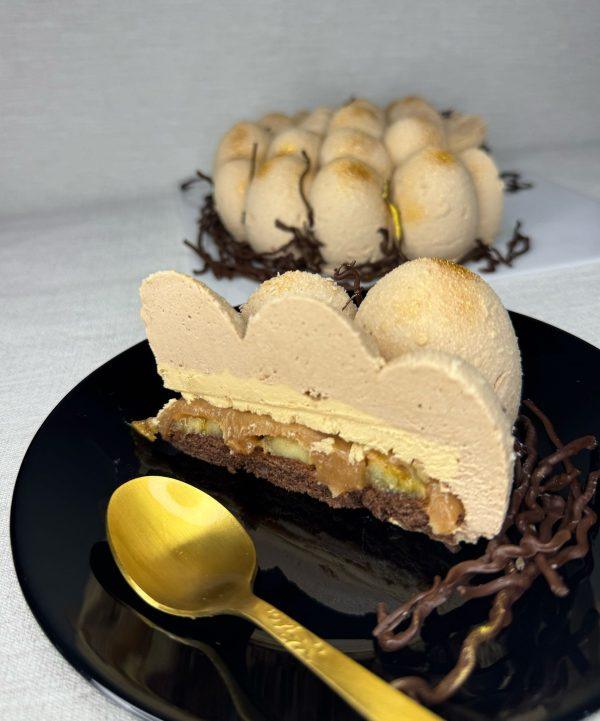 A slice of a layered cake designed to look like an egg from a bird's nest cake, resting on a black plate with a gold spoon. The cake has a light brown mousse exterior, a lighter filling with visible pieces, and a dark base. A whole bird's nest cake is blurred in the background.