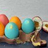 Six colorful, edible Easter eggs (pink, orange, teal) in a broken chocolate carton. Two eggs are cut open, showing layers of sponge, fruit filling, and white ganache by Et Voila! Delicacy